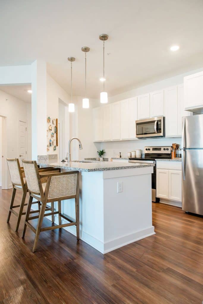 Apartments for rent in Ocala Modern kitchen with white cabinets, stainless steel appliances, a central island with a sink, granite countertops, and two wooden barstools on wooden flooring. Three pendant lights hang above the island. Aurora St. Leon Apartments in Ocala 2150 NW 21st Avenue | Ocala, FL 34475 (352) 233-4133