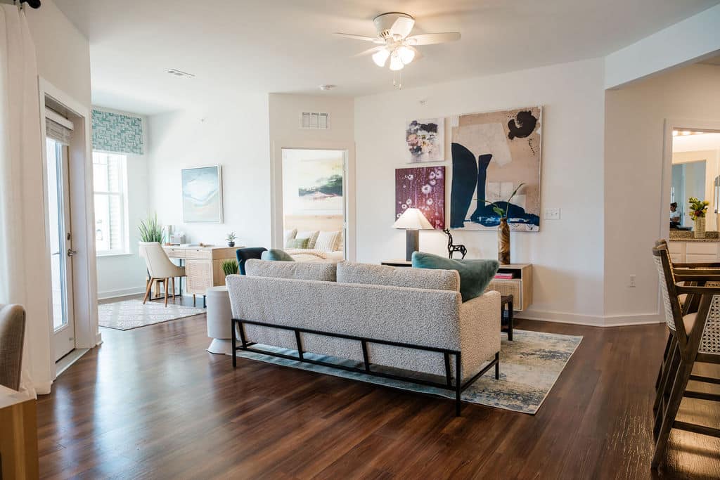 Apartments for rent in Ocala A modern living room with wood flooring, a ceiling fan, and contemporary artwork. It features a beige sofa, two green chairs, a small white side table, and a view into a dining area and kitchen. Aurora St. Leon Apartments in Ocala 2150 NW 21st Avenue | Ocala, FL 34475 (352) 233-4133