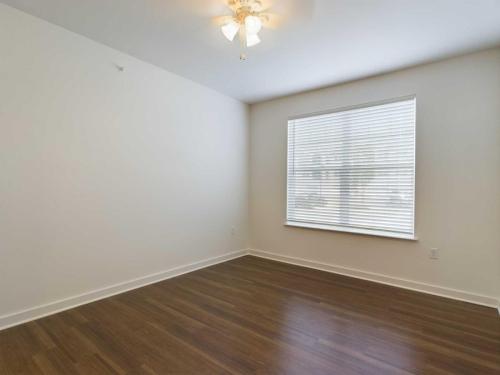 Apartments for rent in Ocala A simple, empty room with white walls and a wooden floor, featuring a ceiling fan with lights and a window with closed blinds allowing some natural light in. Aurora St. Leon Apartments in Ocala 2150 NW 21st Avenue | Ocala, FL 34475 (352) 233-4133