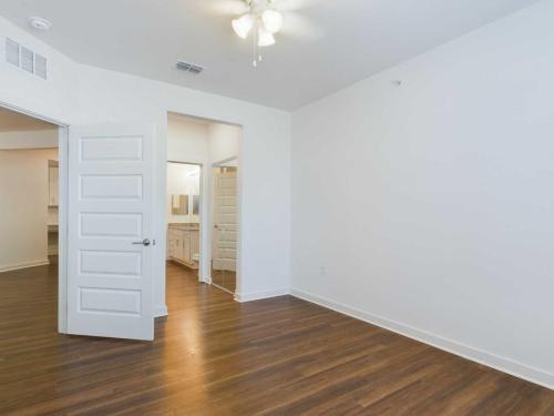 Apartments for rent in Ocala A clean, empty room with white walls, wooden floors, a ceiling fan, an open door to a hallway, and a view of a bathroom with a double sink vanity. Aurora St. Leon Apartments in Ocala 2150 NW 21st Avenue | Ocala, FL 34475 (352) 233-4133