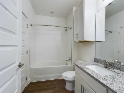 Apartments for rent in Ocala A modern bathroom with a white bathtub, toilet, white cabinets, granite countertop, large mirror, and wood flooring. Aurora St. Leon Apartments in Ocala 2150 NW 21st Avenue | Ocala, FL 34475 (352) 233-4133