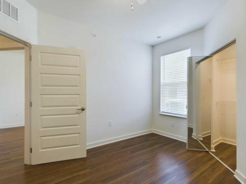 Apartments for rent in Ocala An empty room with wooden floors, an open door leading to another room, a window with blinds, and an open closet with a mirrored door. Aurora St. Leon Apartments in Ocala 2150 NW 21st Avenue | Ocala, FL 34475 (352) 233-4133