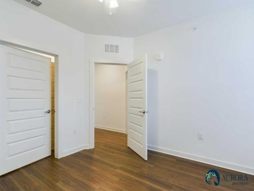 Apartments for rent in Ocala A clean, empty room with white walls, a ceiling fan, wooden flooring, and two open doors. A logo for Aurora Apartments is in the bottom right corner. Aurora St. Leon Apartments in Ocala 2150 NW 21st Avenue | Ocala, FL 34475 (352) 233-4133