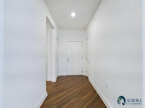 Apartments for rent in Ocala A hallway with white walls, wooden flooring, a recessed ceiling light, and a white door at the end. The logo for Aurora Studio Apartments is at the bottom right. Aurora St. Leon Apartments in Ocala 2150 NW 21st Avenue | Ocala, FL 34475 (352) 233-4133