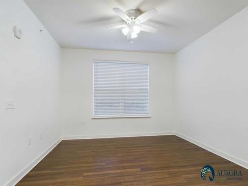 Apartments for rent in Ocala A bright, empty room with white walls, a window with blinds, a ceiling fan, and dark hardwood floors. The Aurora Real Apartments logo is in the bottom right corner. Aurora St. Leon Apartments in Ocala 2150 NW 21st Avenue | Ocala, FL 34475 (352) 233-4133