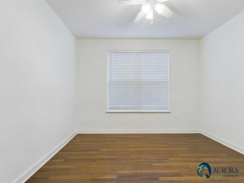 Apartments for rent in Ocala Empty room with white walls, wooden flooring, a closed window with blinds, and a ceiling fan. The Aurora Apartments logo is visible in the bottom right corner. Aurora St. Leon Apartments in Ocala 2150 NW 21st Avenue | Ocala, FL 34475 (352) 233-4133