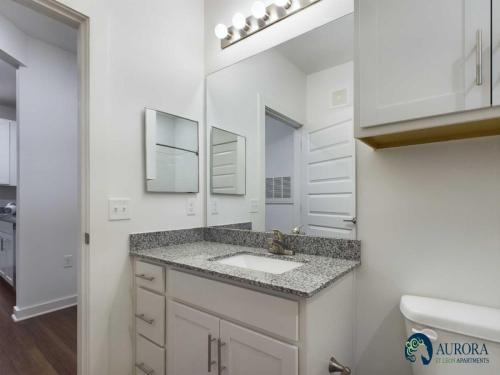 Apartments for rent in Ocala Modern bathroom with granite countertop, mirror, and white cabinets under bright lighting. Door leads to adjacent room. Aurora St. Leon Apartments logo visible. Aurora St. Leon Apartments in Ocala 2150 NW 21st Avenue | Ocala, FL 34475 (352) 233-4133