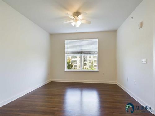 Apartments for rent in Ocala A bright, empty room with hardwood floors, a ceiling fan, a large window with blinds, and white walls. The Aurora Yellow Apartments logo is in the bottom right corner. Aurora St. Leon Apartments in Ocala 2150 NW 21st Avenue | Ocala, FL 34475 (352) 233-4133