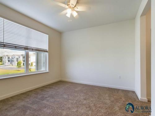 Apartments for rent in Ocala A vacant room with beige carpet, a ceiling fan, large window with blinds, and walls painted white. The Aurora Apartments logo is in the bottom right corner. Aurora St. Leon Apartments in Ocala 2150 NW 21st Avenue | Ocala, FL 34475 (352) 233-4133