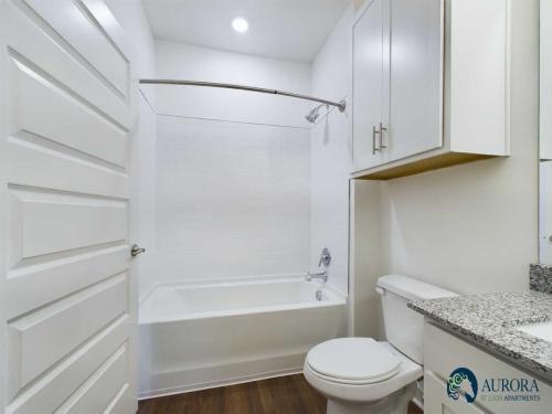 Apartments for rent in Ocala A clean, white bathroom with a bathtub, curved shower rod, toilet, and sink with granite countertop, featuring wooden flooring and white cabinets. Aurora St. Leon Apartments in Ocala 2150 NW 21st Avenue | Ocala, FL 34475 (352) 233-4133