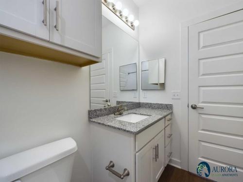 Apartments for rent in Ocala Modern bathroom with white cabinets, granite countertop, undermount sink, large mirror, and overhead vanity lights. A closed door and toilet are also visible. Aurora Stelon Apartments logo in the corner. Aurora St. Leon Apartments in Ocala 2150 NW 21st Avenue | Ocala, FL 34475 (352) 233-4133