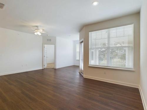 Apartments for rent in Ocala A bright, empty room with wooden flooring, a large window with blinds, a ceiling fan, and a doorway leading to another room. Aurora St. Leon Apartments in Ocala 2150 NW 21st Avenue | Ocala, FL 34475 (352) 233-4133