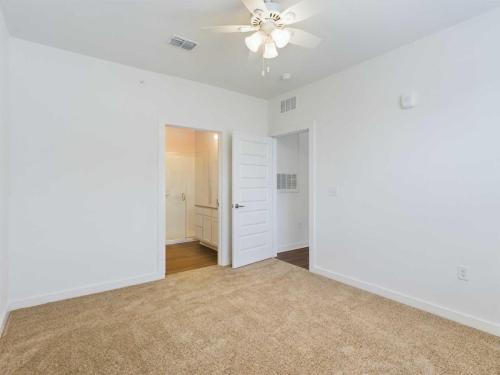 Apartments for rent in Ocala A brightly lit empty bedroom with beige carpeting, white walls, a ceiling fan, an open door leading to a bathroom, and another closed door. Aurora St. Leon Apartments in Ocala 2150 NW 21st Avenue | Ocala, FL 34475 (352) 233-4133