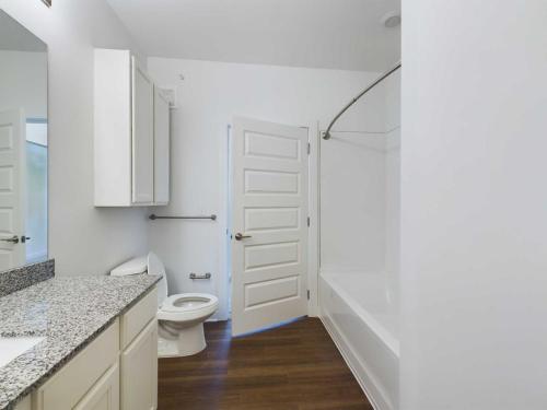 Apartments for rent in Ocala A clean, modern bathroom with a white vanity, granite countertop, bathtub with a shower curtain rod, toilet, and hardwood flooring, featuring a partially open door. Aurora St. Leon Apartments in Ocala 2150 NW 21st Avenue | Ocala, FL 34475 (352) 233-4133