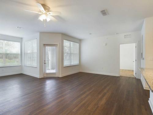 Apartments for rent in Ocala A spacious, empty room with wooden floors, white walls, large windows, a glass door, and a ceiling fan with lights. An adjacent room with a different flooring is visible through an open doorway. Aurora St. Leon Apartments in Ocala 2150 NW 21st Avenue | Ocala, FL 34475 (352) 233-4133