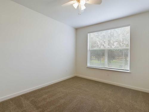 Apartments for rent in Ocala An empty room with beige carpet, white walls, a ceiling fan with light, and a large window with blinds partially open. Aurora St. Leon Apartments in Ocala 2150 NW 21st Avenue | Ocala, FL 34475 (352) 233-4133