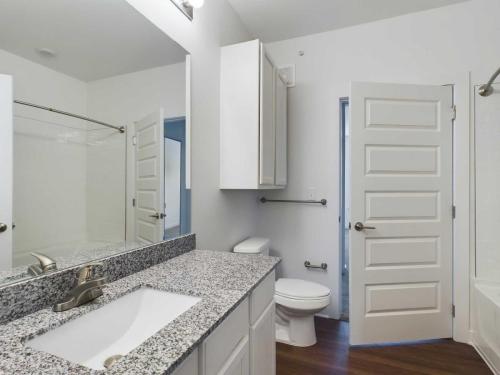 Apartments for rent in Ocala Modern bathroom with a granite countertop sink, toilet, and bathtub. White cabinetry, mirror above the sink, brown floor, and white doors. Aurora St. Leon Apartments in Ocala 2150 NW 21st Avenue | Ocala, FL 34475 (352) 233-4133
