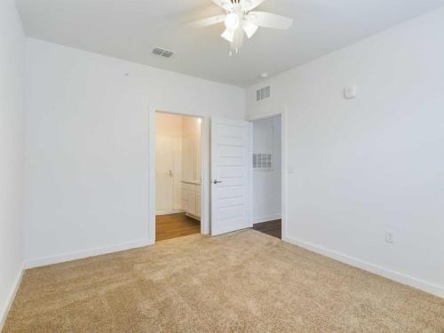Apartments for rent in Ocala A clean, empty room with beige carpet, white walls, a ceiling fan, and an open door leading to another room with hardwood flooring. Aurora St. Leon Apartments in Ocala 2150 NW 21st Avenue | Ocala, FL 34475 (352) 233-4133
