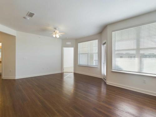Apartments for rent in Ocala An empty room with large windows, hardwood floors, white walls, a ceiling fan, and an open door leading to another room. Aurora St. Leon Apartments in Ocala 2150 NW 21st Avenue | Ocala, FL 34475 (352) 233-4133