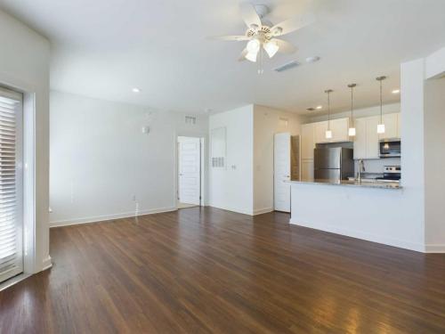 Apartments for rent in Ocala Bright, empty living room with wooden flooring, a ceiling fan, and modern kitchen with stainless steel appliances and pendant lighting in the background. Aurora St. Leon Apartments in Ocala 2150 NW 21st Avenue | Ocala, FL 34475 (352) 233-4133