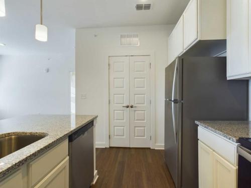 Apartments for rent in Ocala Modern kitchen with granite countertops, stainless steel refrigerator, dishwasher, and white cabinets. Wood flooring and recessed lighting are visible. A double-door pantry is centered in the background. Aurora St. Leon Apartments in Ocala 2150 NW 21st Avenue | Ocala, FL 34475 (352) 233-4133