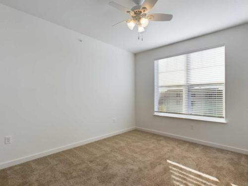 Apartments for rent in Ocala Empty room with white walls, beige carpet, a ceiling fan with lights, and a large window with blinds allowing natural light to enter. Aurora St. Leon Apartments in Ocala 2150 NW 21st Avenue | Ocala, FL 34475 (352) 233-4133