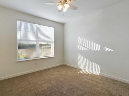 Apartments for rent in Ocala A room with beige carpet, white walls, a ceiling fan, and a large window with closed blinds casting shadows on the wall. Aurora St. Leon Apartments in Ocala 2150 NW 21st Avenue | Ocala, FL 34475 (352) 233-4133