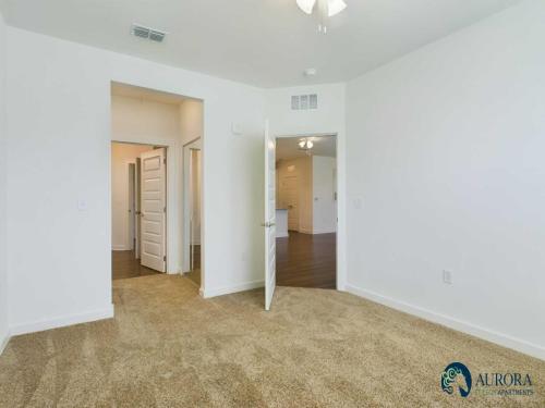 Apartments for rent in Ocala A carpeted, empty room with white walls, an open door leading to a hallway with multiple doors, and a ceiling fan light fixture. Aurora Apartments logo in bottom right corner. Aurora St. Leon Apartments in Ocala 2150 NW 21st Avenue | Ocala, FL 34475 (352) 233-4133
