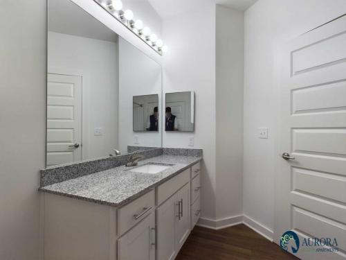 Apartments for rent in Ocala Modern bathroom with single granite countertop sink, dual mirrors, white cabinets, overhead light fixture, and wooden flooring. Aurora Stello Apartments logo visible in bottom right corner. Aurora St. Leon Apartments in Ocala 2150 NW 21st Avenue | Ocala, FL 34475 (352) 233-4133