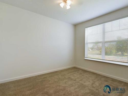 Apartments for rent in Ocala A small, empty room with beige carpet, white walls, a ceiling fan with lights, and a window with closed blinds. The Aurora Apartments logo is visible in the bottom right corner. Aurora St. Leon Apartments in Ocala 2150 NW 21st Avenue | Ocala, FL 34475 (352) 233-4133