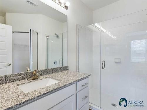 Apartments for rent in Ocala A modern bathroom with a granite countertop, sink, mirror, glass-enclosed shower, and a door leading to a closet. Logo of Aurora St. Leon Apartments in the bottom right corner. Aurora St. Leon Apartments in Ocala 2150 NW 21st Avenue | Ocala, FL 34475 (352) 233-4133