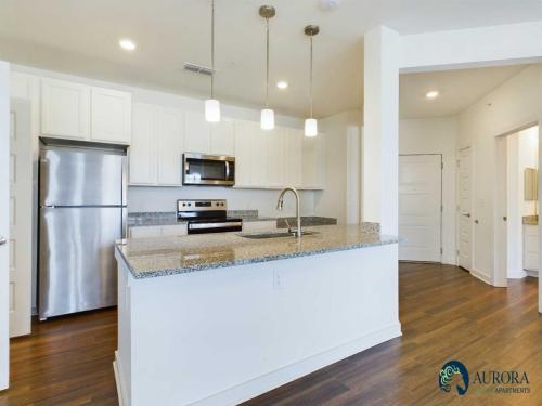 Apartments for rent in Ocala A modern kitchen with stainless steel appliances, granite countertops, white cabinetry, and hardwood floors. The space includes an island with a sink and three overhead pendant lights. Logo: Aurora Apartments. Aurora St. Leon Apartments in Ocala 2150 NW 21st Avenue | Ocala, FL 34475 (352) 233-4133
