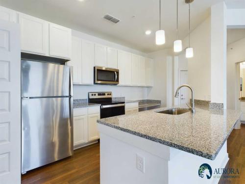 Apartments for rent in Ocala Modern kitchen with stainless steel appliances, granite countertops, white cabinetry, and pendant lighting over a central island. Aurora Steel Apartments logo displayed in the bottom right corner. Aurora St. Leon Apartments in Ocala 2150 NW 21st Avenue | Ocala, FL 34475 (352) 233-4133