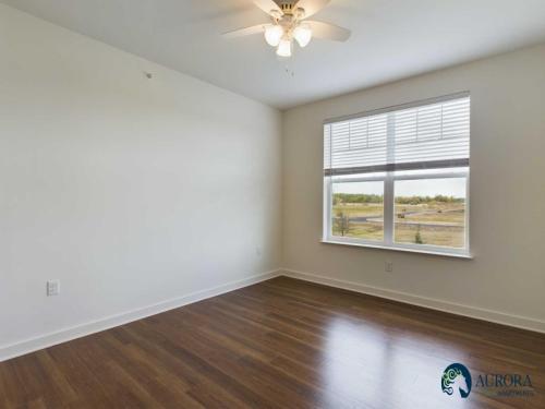 Apartments for rent in Ocala A bright, empty room with wooden floor, white walls, a ceiling fan-light, and a large window overlooking a landscape. The Aurora Apartments logo is in the bottom right corner. Aurora St. Leon Apartments in Ocala 2150 NW 21st Avenue | Ocala, FL 34475 (352) 233-4133