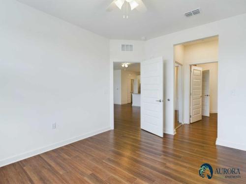 Apartments for rent in Ocala A bright room with white walls, a ceiling fan, and brown hardwood floors. An open door leads to a hallway. The logo in the corner reads "Aurora Apartments. Aurora St. Leon Apartments in Ocala 2150 NW 21st Avenue | Ocala, FL 34475 (352) 233-4133
