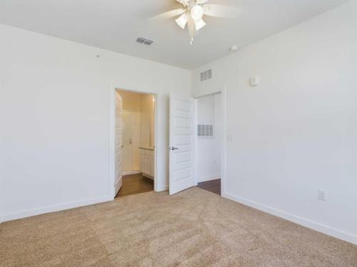 Apartments for rent in Ocala An empty room with beige carpet, white walls, and a white ceiling fan. The door is open, revealing an adjacent room with hardwood flooring and a bathroom. Aurora St. Leon Apartments in Ocala 2150 NW 21st Avenue | Ocala, FL 34475 (352) 233-4133