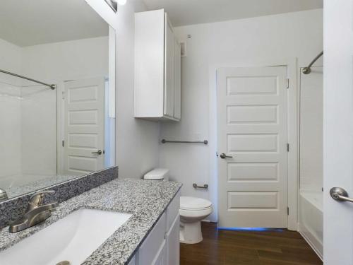 Apartments for rent in Ocala A modern bathroom with a granite countertop, white sink, white cabinets, a toilet, a shower with a white curtain, wood floor, and white doors. Aurora St. Leon Apartments in Ocala 2150 NW 21st Avenue | Ocala, FL 34475 (352) 233-4133