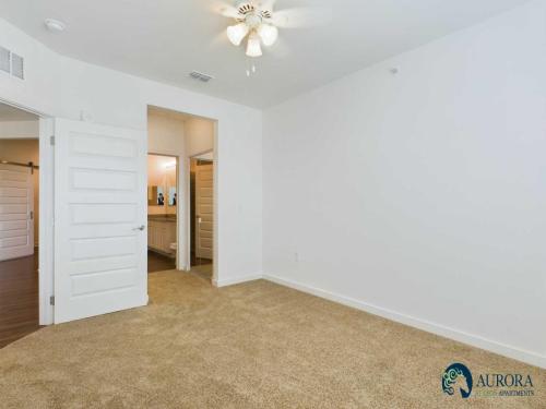 Apartments for rent in Ocala Empty, carpeted room with white walls, a ceiling fan, and a door leading to an adjacent bathroom area. Logo in the bottom right corner reads "Aurora Apartments. Aurora St. Leon Apartments in Ocala 2150 NW 21st Avenue | Ocala, FL 34475 (352) 233-4133