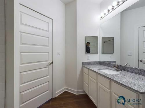 Apartments for rent in Ocala A bathroom with a double sink vanity, large mirror, white cabinets, granite countertop, wooden floor, and a door on the left. Logo in the lower right corner reads "AURORA APARTMENTS. Aurora St. Leon Apartments in Ocala 2150 NW 21st Avenue | Ocala, FL 34475 (352) 233-4133