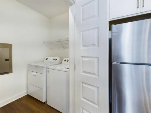 Apartments for rent in Ocala A compact laundry room with a washer and dryer next to a stainless steel refrigerator. The space also includes a white door and a wire shelf above the appliances. Aurora St. Leon Apartments in Ocala 2150 NW 21st Avenue | Ocala, FL 34475 (352) 233-4133