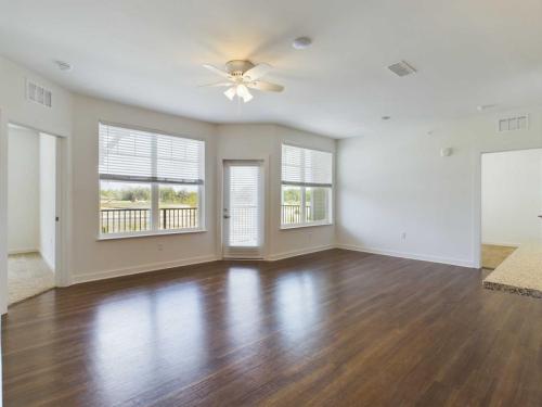 Apartments for rent in Ocala A spacious, empty room with hardwood floors, large windows with blinds, a ceiling fan, and a door leading to a balcony. Two adjoining rooms are visible through open doorways. Aurora St. Leon Apartments in Ocala 2150 NW 21st Avenue | Ocala, FL 34475 (352) 233-4133