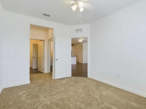Apartments for rent in Ocala A clean, empty room with beige carpet, white walls, a ceiling fan, and two open doors leading to a hallway with hardwood flooring and adjoining spaces. Aurora St. Leon Apartments in Ocala 2150 NW 21st Avenue | Ocala, FL 34475 (352) 233-4133