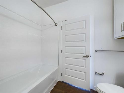 Apartments for rent in Ocala Modern bathroom with a white bathtub, curved shower rod, white tiled walls, wooden flooring, and a white paneled door. The toilet and a small cabinet are visible on the right side. Aurora St. Leon Apartments in Ocala 2150 NW 21st Avenue | Ocala, FL 34475 (352) 233-4133